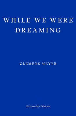 Paperback of "While We Were Dreaming" by Fitzcarraldo Editions, featuring the book cover with abstract artwork and title text.